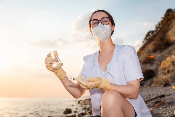 Portrait of young woman scientist-ecologist wearing eyeglasses, mask and rubber gloves examines flask of water from ocean. Copy space. Concept of ecology and science.