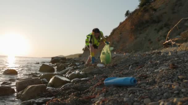 女性ボランティアの低い角度のビューは完全なプラスチックごみ袋を保持し 野生の小石の海岸を歩いてプラスチック製のボトルを拾います 環境汚染の概念 — ストック動画