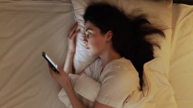 Uyku vakti. Beyaz tenli genç bir kadının yatakta yatarken akıllı telefon kullanmasının en iyi görüntüsü. Uykusuzluk ve sosyal medya bağımlılığı kavramı