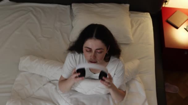 かわいいコーカサス人の若い女性のトップビューはベッドに横たわり 睡眠のためにリラックスした音楽にヘッドフォンとリストを着ています スマートガジェットとテクノロジーの概念 — ストック動画