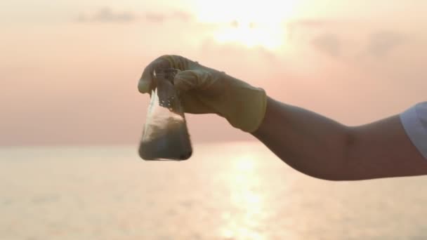 在橡胶手套测试样品瓶的脏水并摇动它的手的近景 背景是日落和大海 水污染与生态概念 — 图库视频影像