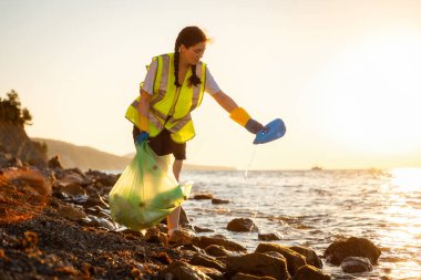 Lastik eldivenli genç beyaz gönüllü kadın vahşi çakıl taşı sahillerinde çöp topluyor. Uzayı kopyala Ekolojik felaket ve temiz okyanus kavramı.