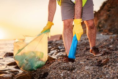 Gönüllü, çakıl taşlı kumsaldan plastik şişe alıyor. Bacaklarını kapat. Çevre koruma kavramı ve Dünya Günü.