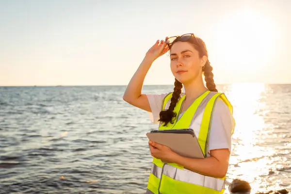 Inspection environmentalist and analyzing water in ocean. Portrait of young volunteer holds tablet wearing vest. In background is ocean, coast and sunset. Concept of Earth Day and ecology.