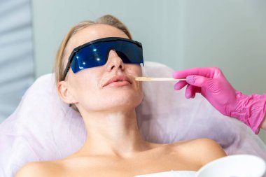 Lazer tüy çıkarma seansında genç bir kadının portresi. Kozmetik uzmanı kadının üst dudağına jel sürüyor. Lazer tüy çıkarma kavramı.