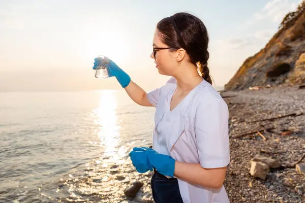 Ecologist wearing protective gloves and eyeglasses, holds research sample flask of water test. Concept of pollution water and environment protection.