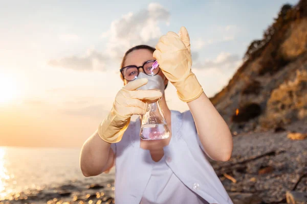 Portrait of young woman scientist-ecologist wearing eyeglasses, mask and rubber gloves examines and tests flask sample of water. Concept of science and laboratory experiments.
