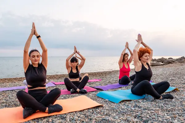 Outdoor activity at sea coast. Side view of group of Caucasian women meditate sitting on sports mats on pebble beach. Yoga asana for meditating.