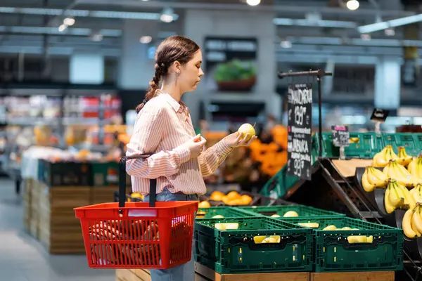 Genç beyaz kadın alışveriş arabasını tutuyor ve süpermarketten meyve seçiyor. Yan görüş. Organik tarım sebze ve meyve alma kavramı.