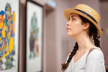 Hasır şapkalı genç beyaz kadın portresi galeride tablolara hayran. Yan görüş. Müzede kültürel eğitim kavramı.