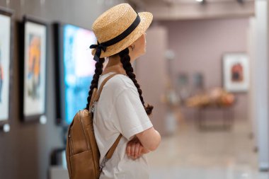 Hasır şapkalı beyaz bir kadının galeride tablolara hayran bakışları. Müze ve kültür eğitimi kavramı.