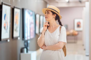 Tarihsel eğitim. Hasır şapka takan güzel beyaz bir kadın galeride tablolara hayran. Müze ve kültür eğitimi kavramı.