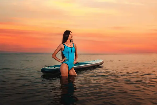 stock image A young slim woman in swimsuit posing with a sup board standing in the water. Sunset in the background and sea. Copy space. Concept of sport and summer activity.