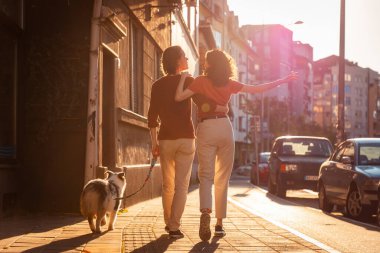Mutlu, kucaklaşan bir çiftin arka planında erkek ve kadın köpekleriyle birlikte şehirde dolaşıyorlar. Arkaplanda gün batımı ışığı.