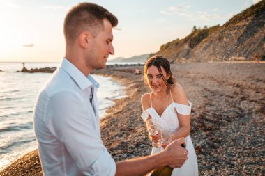 The Wedding day. Husband and wife cheerfully open a bottle of champagne. The ocean is in the background. Newlyweds.