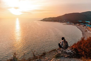 Turizm ve spor eğlencesi. Sırtında turist çantası olan genç bir kadın bir kayanın üzerinde oturur ve gün batımını ve denizi seyreder. Körfez manzarası. Boşluğu kopyala.