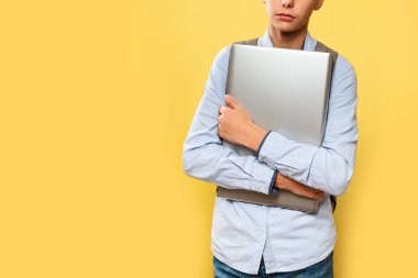 Modern teknoloji ve eğitim kavramı. Mavi gömlekli ve sırt çantalı beyaz bir çocuk dizüstü bilgisayarını kavradı. Sarı arka plan. Boşluğu kopyala ve kapat.