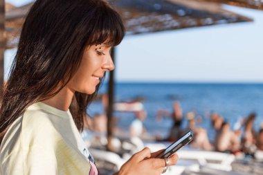 Genç bir kadın telefon ekranına bakıyor. Arka planda insanlarla dolu bir deniz plajı var. İnternet ve sosyal ağ üzerinde uzaktan çalışma kavramı.