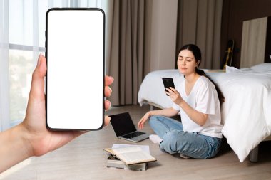 Yerde oturan ve akıllı telefon kullanan genç bir kadının portresi. Bir dişinin eli akıllı telefonu tutar. Yerde bir kitap ve dizüstü bilgisayar var. Model yap. Çevrimiçi öğrenme ve kurye kavramı.