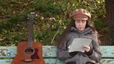 Güzel beyaz bir kadının ortalama planı elinde gitarla bankta oturup not defterinde notlar çalışmak. Müzik aletleri çalmayı öğrenmek.