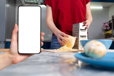 Bir dişinin eli akıllı telefonu maketle tutuyor. Arka planda, bir kadın rendeleyiciye peynir sürer. Modern teknoloji ve yemek pişirme kavramı.