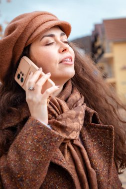 Diş teli, bere ve palto takmış beyaz, mutlu bir kadının portresi sokaktaki akıllı telefondan aranır. Modern sosyal medya kavramı.