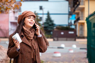 Bereli ve ceketli genç beyaz bir kadının portresi caddedeki akıllı telefondan aranmış ve elinde bir fincan kahve tutuyormuş. Modern sosyal medya kavramı.