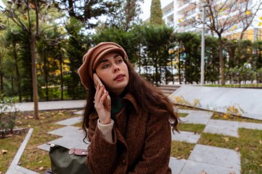 Şapka ve palto giymiş, telefonda konuşan, sonbahar parkında oturan beyaz bir kadının orta pozu. Modern iletişim ve iş
