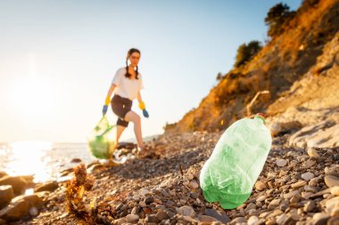 Plastik eldivenli gönüllü bir kadın plastik torba dolusu çöpü tutuyor ve çakıl taşlı vahşi plajda yürüyor. Ön plandaki plastik şişeyi kapat..