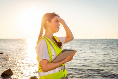 Okyanustaki suyu inceleyen çevreci biri. Tablet tutan genç gönüllülerin yan görüntüsü. Arka planda okyanus, sahil ve günbatımı var. Çevre koruma kavramı.