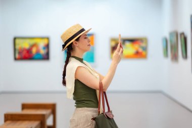 Zarif takım elbiseli beyaz bir kadının yan görüntüsü bir sanat galerisinde ve internet gezisi için akıllı telefonunu kullanıyor. Modern kültür eğitimi müzede.