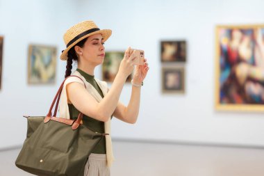 Zarif giysiler ve hasır şapkalı beyaz bir kadının sanat galerisinde akıllı telefonuyla fotoğraf çektiği orta resim. Yan görüş. Modern kültür eğitimi ve çevrimiçi gezi.