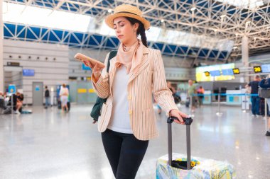 Zarif takım elbiseli güzel beyaz bir kadının orta pozu akıllı telefon kullanıyor ve bavul tutuyor. Havaalanında biri var. Seyahat kavramı ve online seyahat uygulaması.