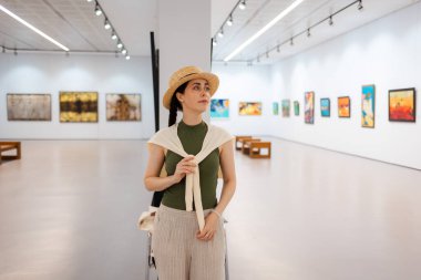 Saman şapkalı, zarif giyimli genç bir kadının galerideki resim sergisine hayran bir portresi. Müzedeki çağdaş sanat eserleri. Kültürel eğitim kavramı.