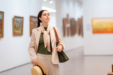 Hasır şapkalı ve takım elbiseli, sanat galerisinde çanta taşıyan ve sanat eserleri tasarlayan yetişkin beyaz bir kadının orta pozu. Kültür Eğitimi ve Müze Günü kavramı.