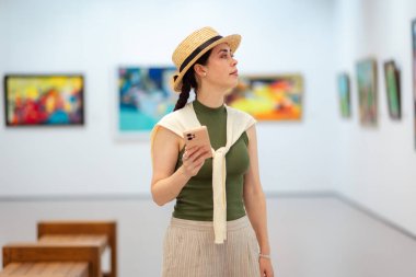 Beyaz tenli güzel bir kadının zarif bir takım elbiseyle bir sanat galerisinde çevrimiçi gezintide akıllı telefonunu kullanırken görüntüsü. Modern kültür eğitimi müzede.