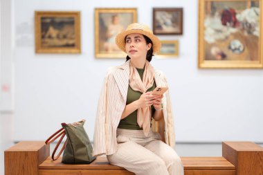 Zarif takım elbiseli beyaz bir kadının sanat galerisindeki bir bankta otururken ve akıllı telefonunu tutarken orta pozu. Modern kültür eğitimi müzede.