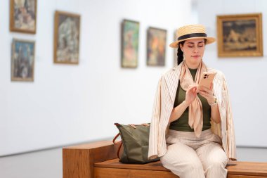 Zarif bir takım elbiseli beyaz bir kadının sanat galerisindeki bir bankta oturup akıllı telefonuyla online bir turu izlediği bir fotoğraf. Uzayı kopyala Modern kültür eğitimi.