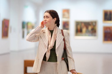 Genç, beyaz, zarif bir kadının, çağdaş bir sanat sergisini ziyaret ederken şüphe ve endişeye kapıldığı bir fotoğraf. Resimler arka planda. Müze günü ve kültür eğitimi kavramı.