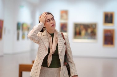 Çağdaş bir sanat sergisini ziyaret ederken genç, beyaz, zarif bir kadının resmi başını tutuyor. Resimler arka planda. Müze günü ve kültür eğitimi kavramı.