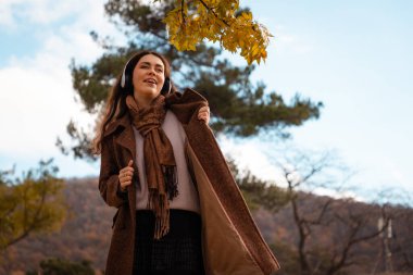 Palto giymiş, kulaklıkla müzik dinleyen ve sonbahar parkında dans eden güzel, mutlu, beyaz bir kadın resmi. Eğlence ve psikoloji kavramı.
