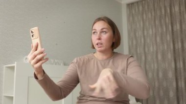 Yetişkin, beyaz bir kadının oturma odasında akıllı telefondan video ile iletişim kurarken görüntüsü. Video çağrısı ve sosyal medya kavramı.
