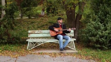 Beyaz sakallı, yakışıklı bir adamın parkta bankta gitar çalarken geniş açılı görüntüsü. Gitar çalmayı öğreniyorum..
