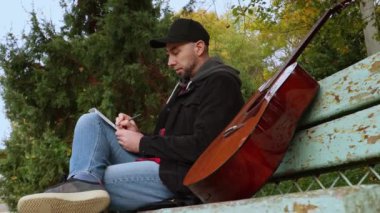 Yetişkin, beyaz bir adamın bankta gitar için müzik bestelemesi. Arkaplana park et