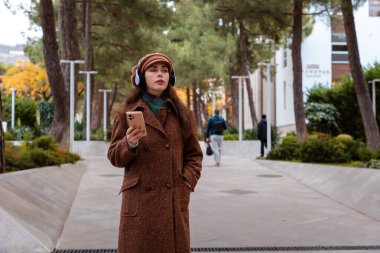 Şapkalı, paltolu, kulaklıklı, müzik dinleyen ve akıllı telefon kullanan güzel beyaz bir kadının orta pozu. Psikoloji kavramı ve rahatlama.