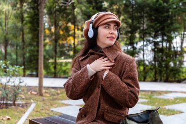 Parkta müzik dinleyen, şapkalı ve kulaklıklı güzel, hayalperest, beyaz bir kadının orta pozu..
