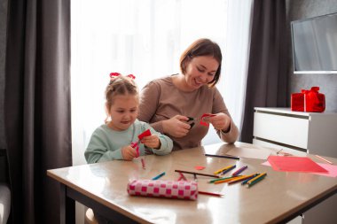 Gülümseyen anaokulu öğretmeni küçük bir kızın Sevgililer Günü için el işi yapmasına yardım ediyor. Küçük bir kız kağıt kalp kesiyor. Anaokulu ve Anneler Günü konsepti.