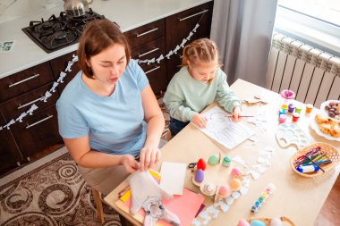 Genç beyaz bir anne ve kızı tavşancık maskesi deseninde süslemişler. Aile mutfak masasında oturmuş şenlik dekorasyonu yapıyor.
