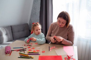 Kafkasyalı genç bir anne ve küçük tatlı kızının masa başında oturan kağıt kalpler çıkardığı bir fotoğraf. Aile Sevgililer Günü kutlaması için hazırlanıyor..