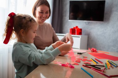 Beyaz kadın anaokulu öğretmeni, küçük tatlı bir kıza Sevgililer Günü 'nde kırmızı kağıtlardan nasıl kalp kesileceğini öğretiyor..